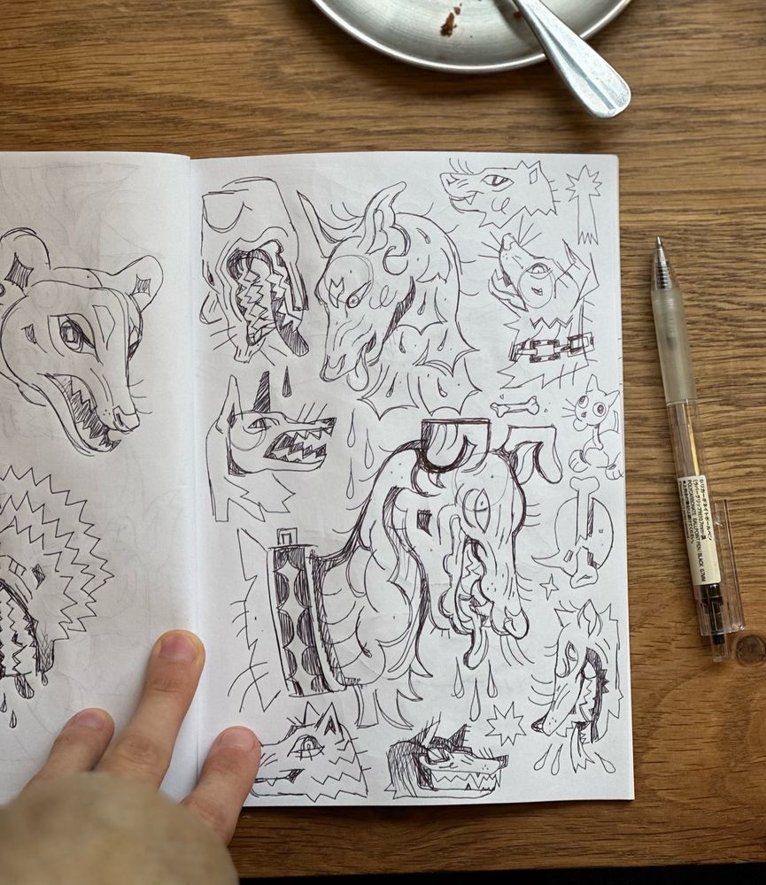 Sketchbook page laying on wooden table with pen next to it. It’s filled with dog-like creatures drawn with black pen.