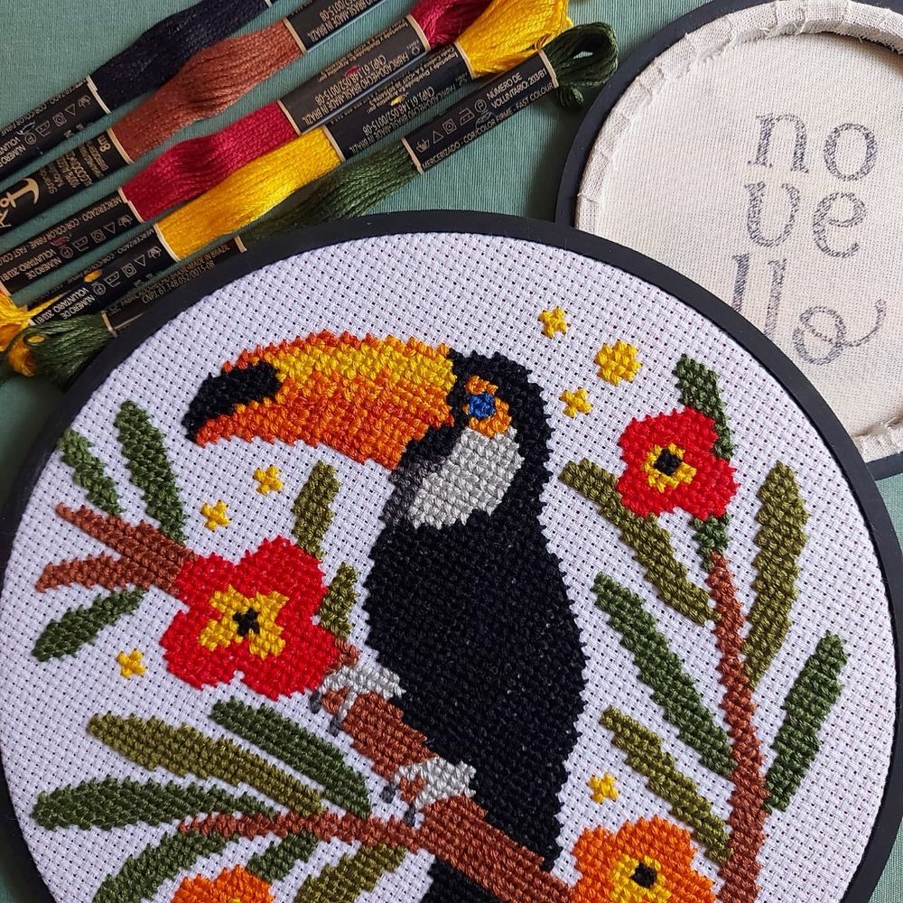 Foto de Bordado em ponto cruz de um tucano em um galho marrom com flores vermelhas e laranjas. Ao fundo, a logo Novello carimbada num tecido