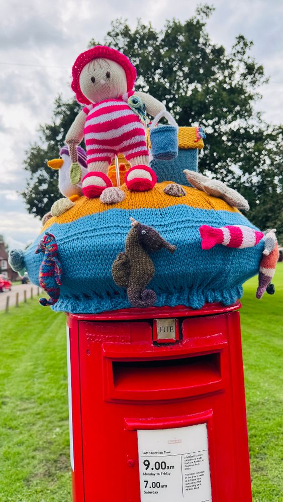 Post box topper on red pillar box with knitted figure in swimming costume, seagull, beach hut and seashells and sea horses 