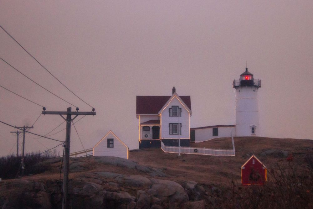 Photo of nubble lighthouse in Maine at dusk. The lighthouse is lined with yellow lights.