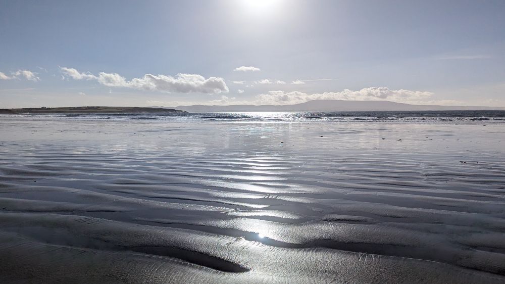 Low level photo across sunlit ripples of sand on a beach just after the tide has gone out. The sea and hills on the far side of a bay are visible on the horizon.