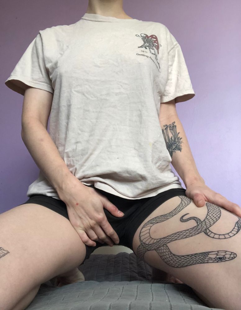 transmasc person in black boxers, a white t shirt, with a snake tattoo on their thigh on their knees facing the camera