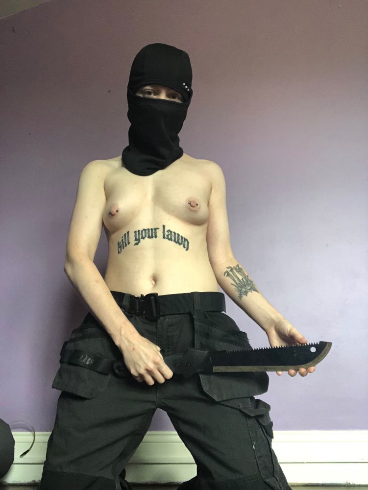 topless transmasc person wearing cargopants and a balaclava, holding a machete and caressing the blade