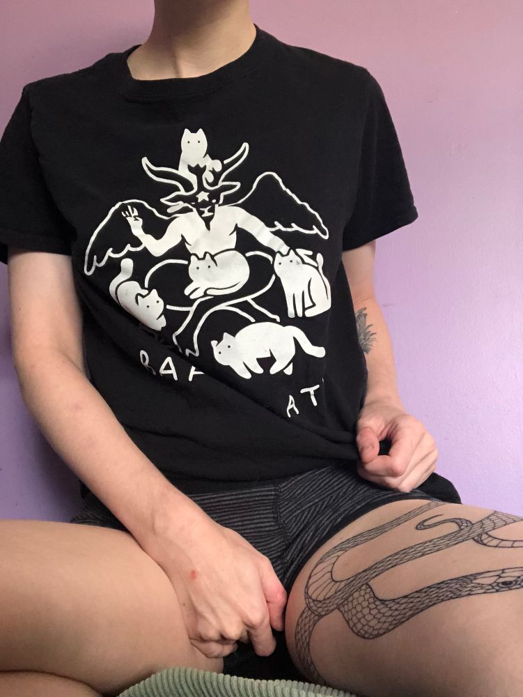 transmasc person sits with legs open toward the camera. They wear boxers and a baphomet/cat shirt, and their right hand is against their crotch