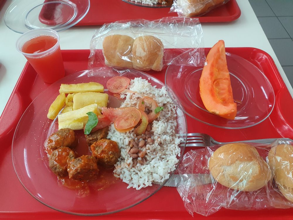 Imagem contendo uma bandeja vermelha e nela, um prato grande com comidas salgadas, um prato menor com a fruta e os pães embalados separadamente. E um copo de refresco de goiaba.