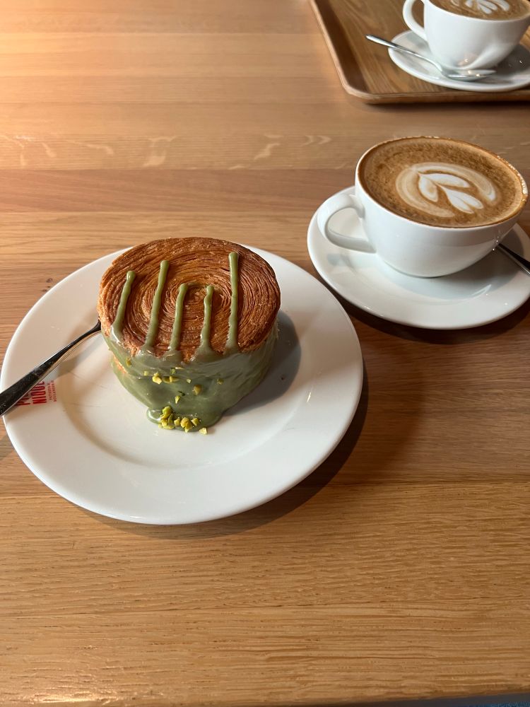 Croissant Roulé Pistache auf einem Teller. 
Daneben ein Cappuccino. Beides auf einem Holztisch 