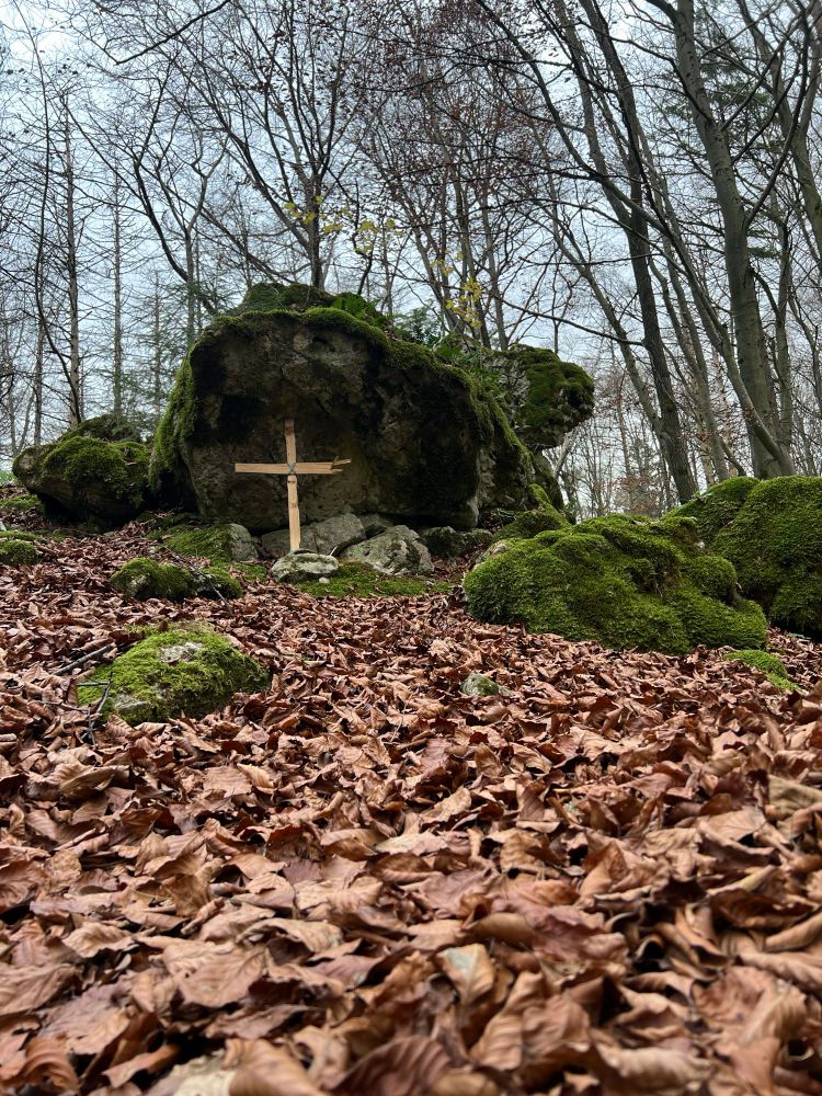 Bemooste Felsen in einem Wald. Am Fuß des Felsen steht ein Holzkreuz. Braunes Laub bedeckt den Boden
