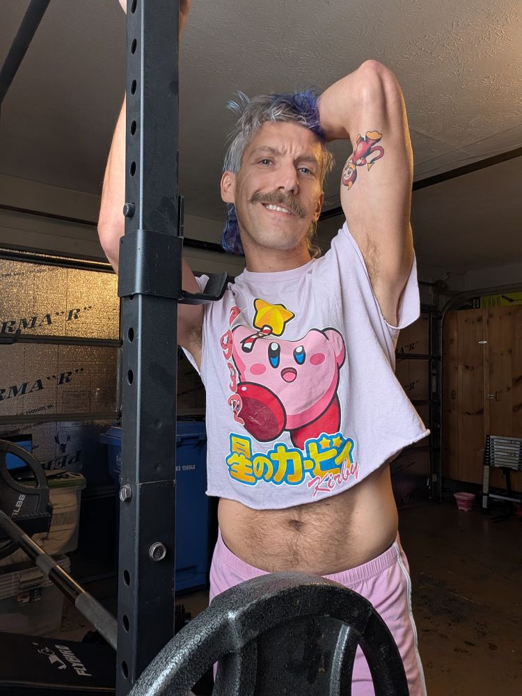 A person in a pink Kirby crop top and shorts leans slightly on a squat rack in a garage gym. They’re smiling gently, with a calm and reflective expression. Gym equipment is visible in the background.