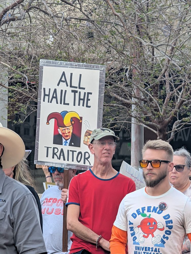 A protester holds up a sign reading "ALL HAIL THE TRAITOR" with an image of Donald Trump wearing a jester hat. A crowd of demonstrators stands around them, some wearing sunglasses and protest-themed t-shirts. In the background, another sign partially visible says "NO ONE IS FREE."

시위자가 "모두 배신자를 찬양하라"라고 적힌 팻말을 들고 있으며, 도널드 트럼프가 어릿광대 모자를 쓴 이미지가 포함되어 있다. 주변에는 선글라스를 끼거나 시위 관련 티셔츠를 입은 시위대가 서 있다. 배경에는 "아무도 자유롭지 않다"라는 문구가 부분적으로 보이는 또 다른 팻말이 있다.

Un manifestante sostiene un cartel que dice "TODOS SALUDEN AL TRAIDOR" con una imagen de Donald Trump usando un sombrero de bufón. A su alrededor, una multitud de manifestantes está reunida, algunos con gafas de sol y camisetas con mensajes de protesta. En el fondo, se ve parcialmente otro cartel que dice "NADIE ES LIBRE."