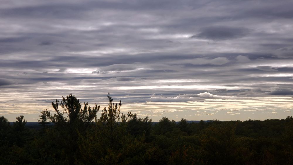 A hilltop view with a distant horizon. The sky is overcast with clouds forming horizontal stripes that appear to stretch almost to the horizon.