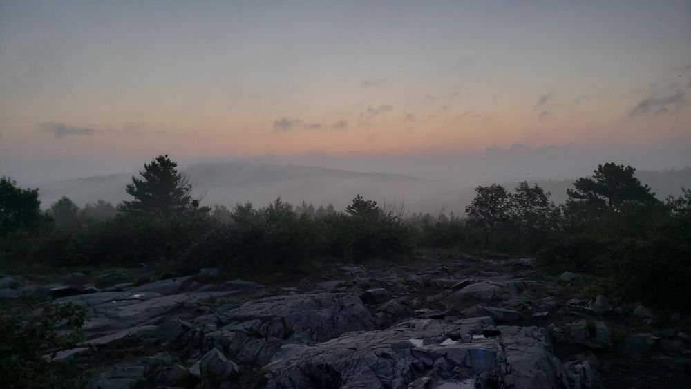 The rocky top of Buck Hill is in the foreground. It is just before sunrise, but low clouds and fog obscure much of the view. Above the clouds a band of orange light gives way to a light blue sky.