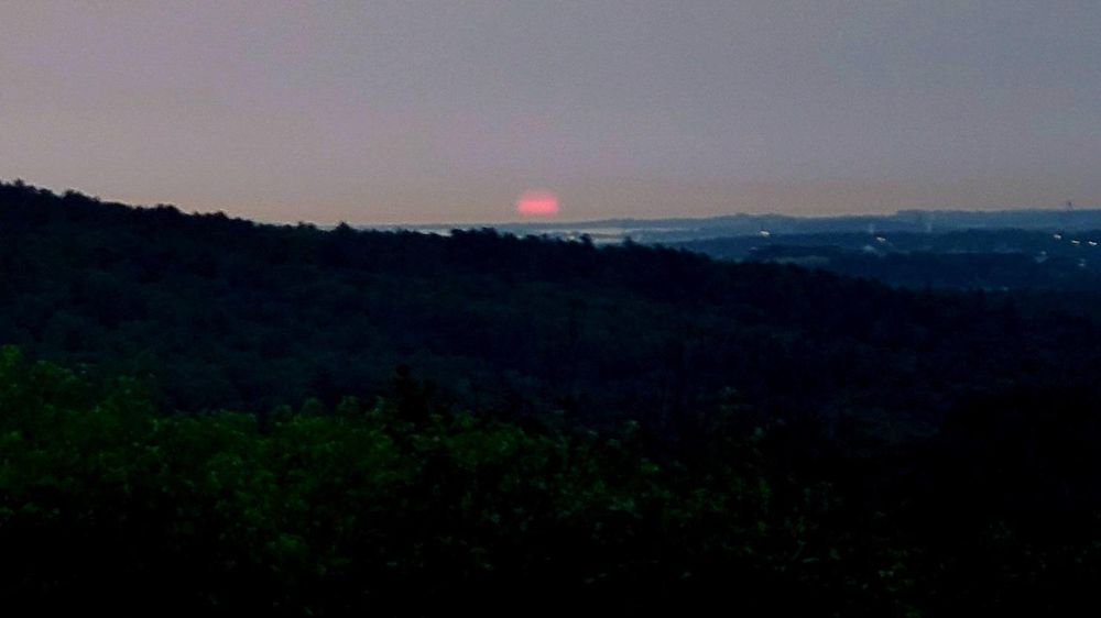 A very red, cloud streaked disc of the sun rises above the horizon as seen from the top of Buck Hill.