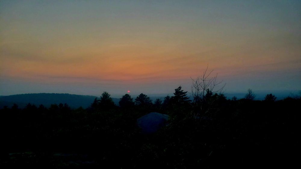 Sunset this evening, June 12th, from Buck Hill! A red disc of the sun sits just above the horizon. Colors of gray, blue and orange paint the sky above the sun.