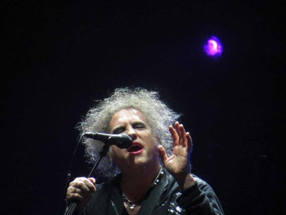 Robert Smith of The Cure singing into a mic under spotlight. 