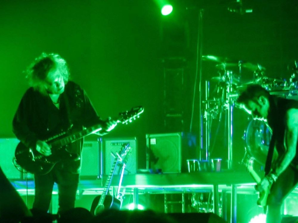 The Cure band members Robert Smith playing guitar and Simon Gallup playing bass under green stage lighting. 