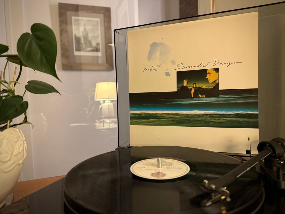 A copy of a-ha’s 1986 album Scoundrel Days is being played on a turntable, with the cover propped up behind. 