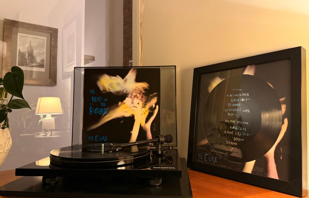 A copy of The Cure record The Head On The Door is playing on a turntable with the album cover propped up behind it. To the side is a framed copy of the picture disc version of the record issued for Record Store Day 2025. The b side is displayed with the track listing pictured in the vinyl disc. 
