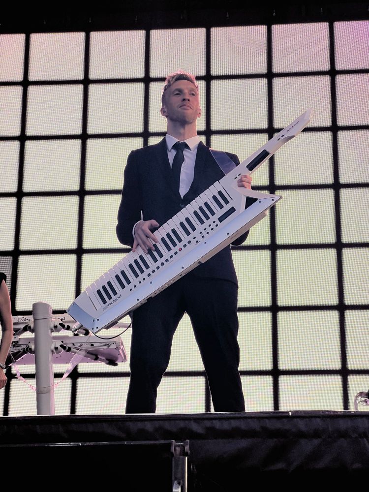 Human lead keyboardist Nick Banks wearing a dark suit and tie, standing on stand and holding a white keytar. 
