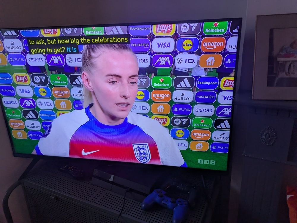 England's Chloe Kelly being interviewed after their Euros win, in front of an advertising hoarding displaying dozens of oblong tiles for different brands.