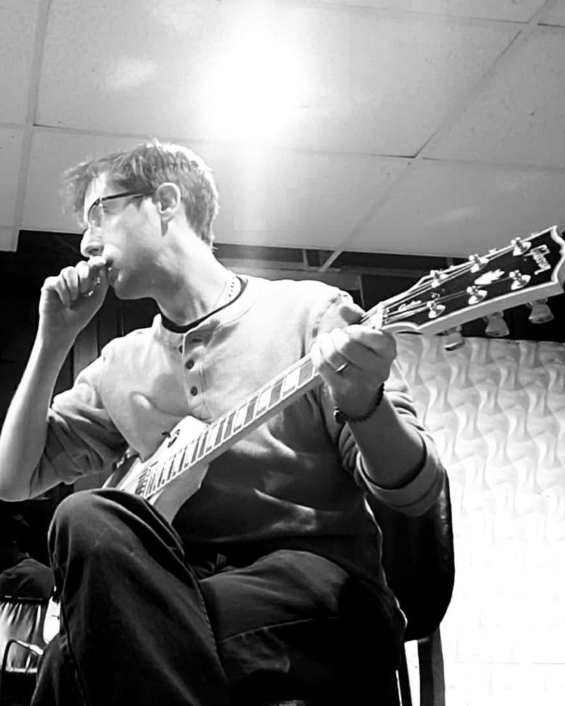 J. Trafford seated in studio with electric guitar.