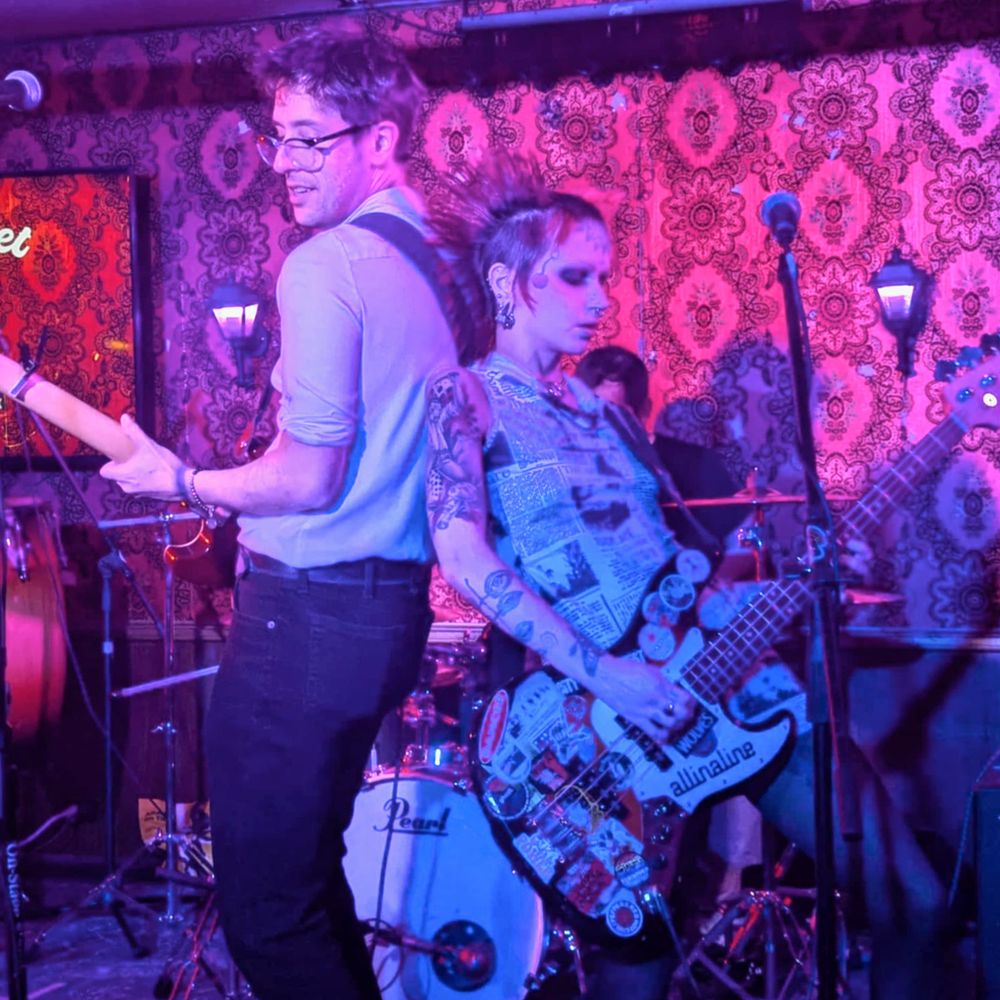 Two people playing guitar and bass respectively, performing back-to-back. There are two microphones in front of them, and behind them a drummer is performing partially obscured.