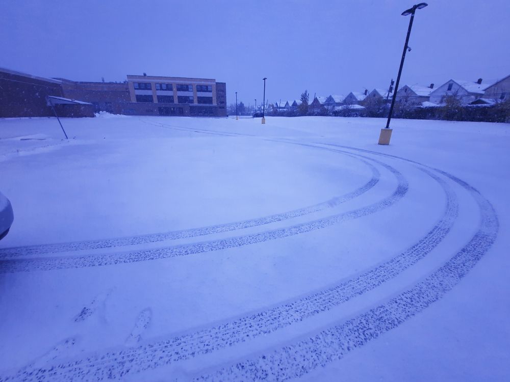 A snow covered parking lot with a single set of tire tracks. A three-story building and a row of houses are in the background 