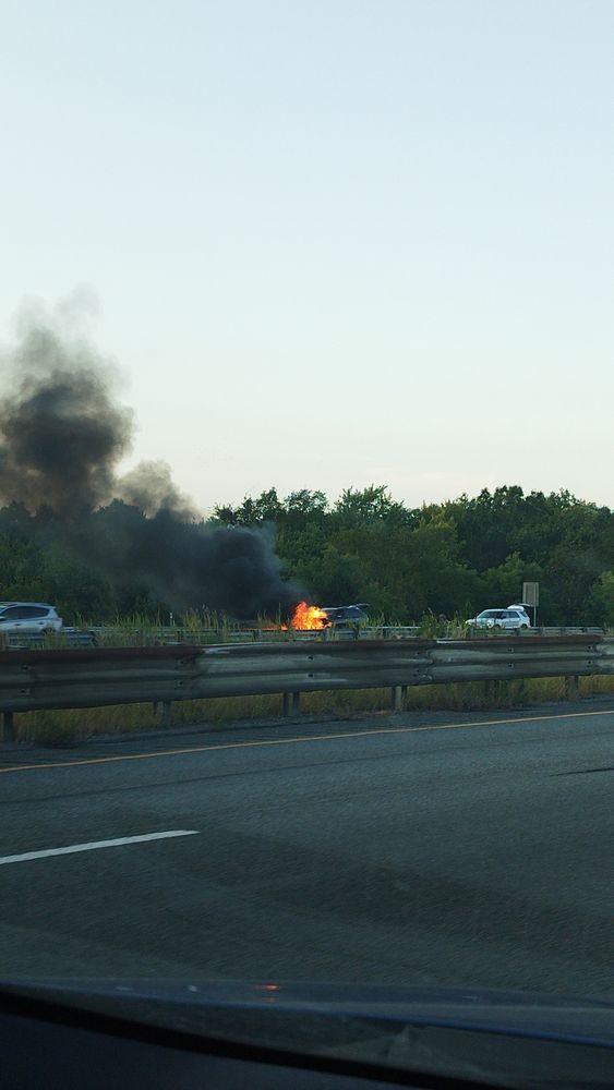 Image of a highway taken from a car. On the opposing lane's shoulder, an SUV is on fire. The fire is coming from the front, thick black smoke blows off into the sky.