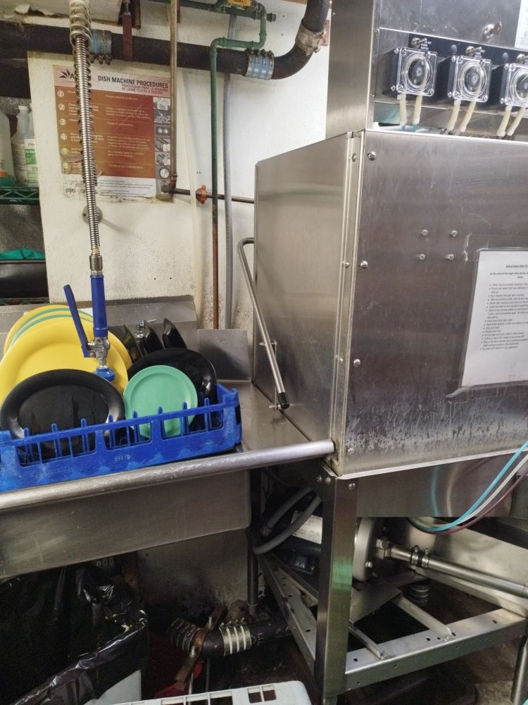 A tray of plates rests on one side of a closed industrial dishwasher. A trash can with a black bag is underneath. 