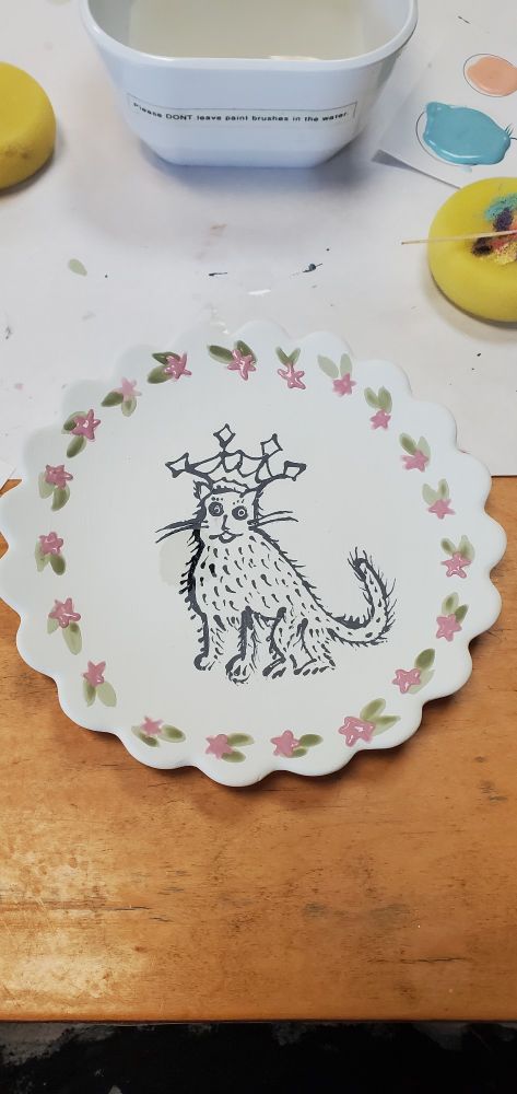 A scalloped dish with a medieval cat painted in the center. The cat is cricled in green leaves and pink flowers. Behind the plate is a light brown wooden table and a white placemat. There are various paint brushes and sponges at the top corners.