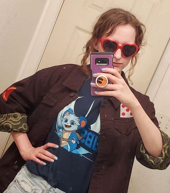 Bluesky user StarrySyndulla looking in a mirror with a neutral expression. She has on a brown and green jacket that resembles Hera Syndulla's in the TV show Ahsoka. She also has on a blue t-shirt with the character Nubs from young jedi adventures on it. She's holding her purple phone case that has Sabine's star bird on it. And lastly she has on red heart sunglasses.