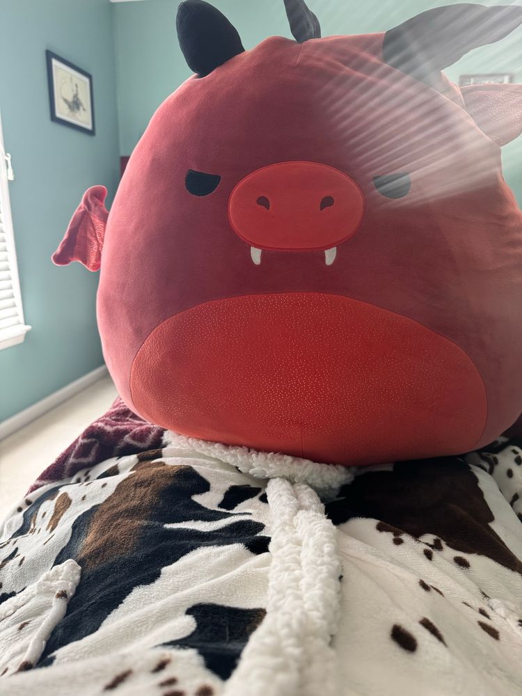 A 24” red dragon squishmellow (big soft stuffed animal) resting on a blanket with a brown/black/white jersey cow pattern on it. 