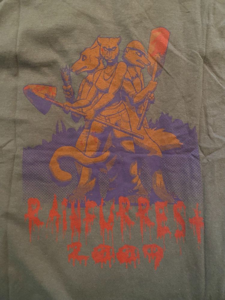 A dark green T-shirt featuring an illustration of three anthropomorphic animals standing back-to-back holding weapons: a wolf with a bloodied broken bottle, a cougar with a bloody shovel, and a weasel wielding a bloody paddle. They are drawn in an orange and purple color scheme, with a forest silhouette behind them. Below the characters, the text "RAINFURREST 2009" is displayed in a red, dripping horror-style font, resembling blood.