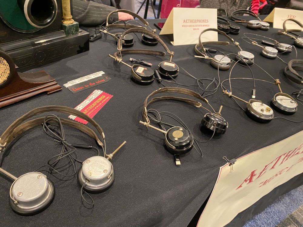 A tabletop display of vintage-style headphones with thin metal headbands and round earpieces, arranged neatly on a black cloth. A sign reads “Aetherphones: Wired and Bluetooth Models - $60.00.” Another vintage speaker device and paper tags that say “Listen to the voices” and “Aetherphones” are visible.