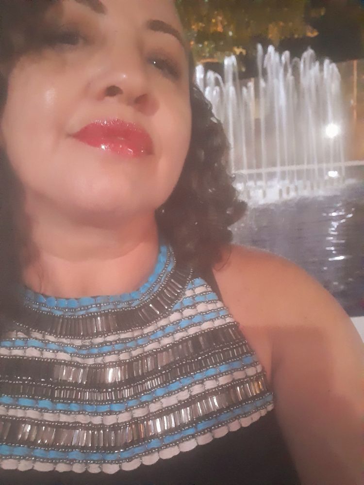 Foto da dona do perfil, mulher parda de cabelo cacheado na altura do ombro, usando blusa preta com um bordado de pedrarias nas cores azul, preto e branco.
Ao fundo tem uma fonte. 
