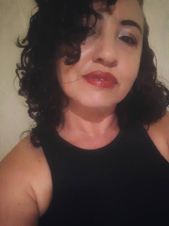 Foto colorida da dona do perfil, mulher parda de cabelo cacheado na altura do ombro, usando blusa preta.