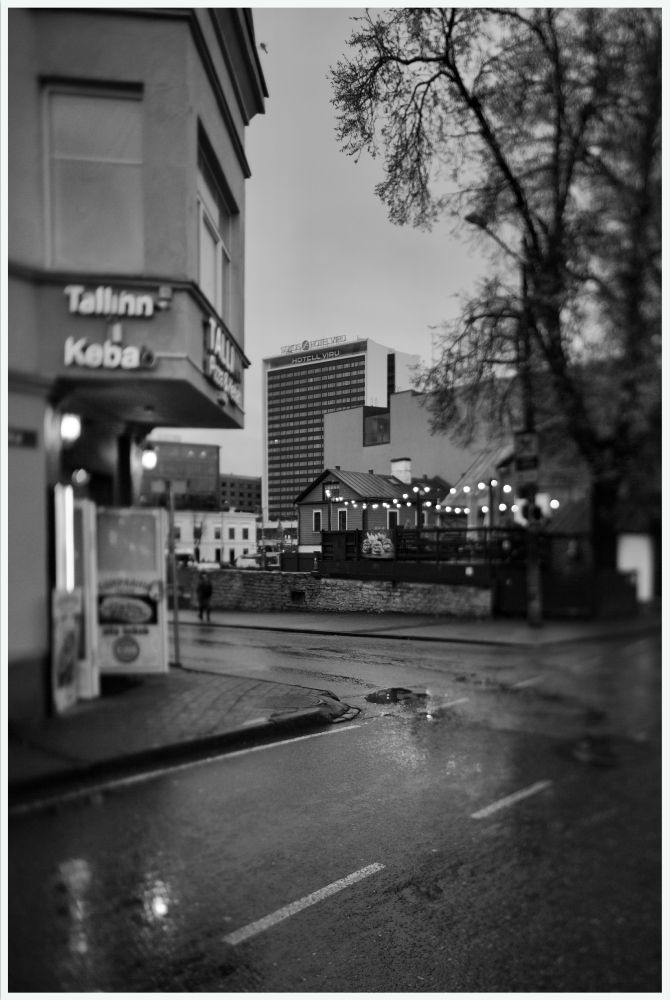 A rainy street in central Tallinn frames the familiar silhouette of Hotel Viru in the distance. The wet pavement and blurred details give it a clean, documentary look of everyday city life.