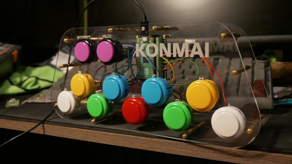 a popn music controller. it is two pieces of clear acrylic with “KONMAI” engraved into it, an intentional misspelling of konami. it has 11 buttons, 9 making up the game with 2 auxiliary buttons. there is a raspberry pi pico in the middle serving as a board. 