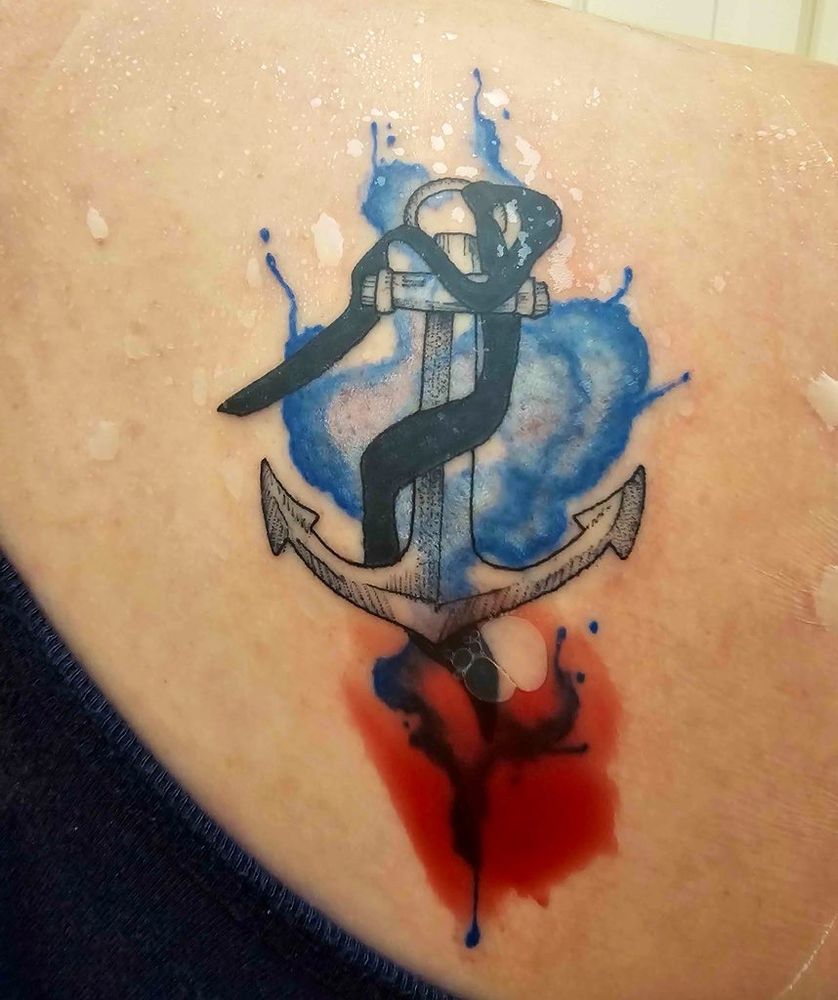 Tattoo of an anchor with a black cravat wrapped around it in place of a rope, with a blue splash watercolour background. The tattoo is wrapped and there is some blood under the covering 