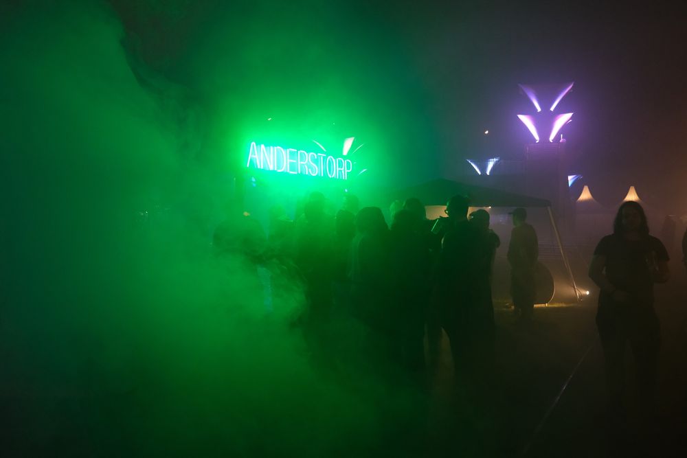 A photo of a very foggy outdoor area. There is a large group of people standing around a tent with a bright green neon light. The fog is moving towards the people 
