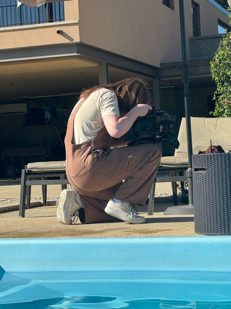 A photo of me recording something with my large video camera. I'm kneeling while balancing the camera on my kee/leg, looking down into the camera to see the viewfinder 