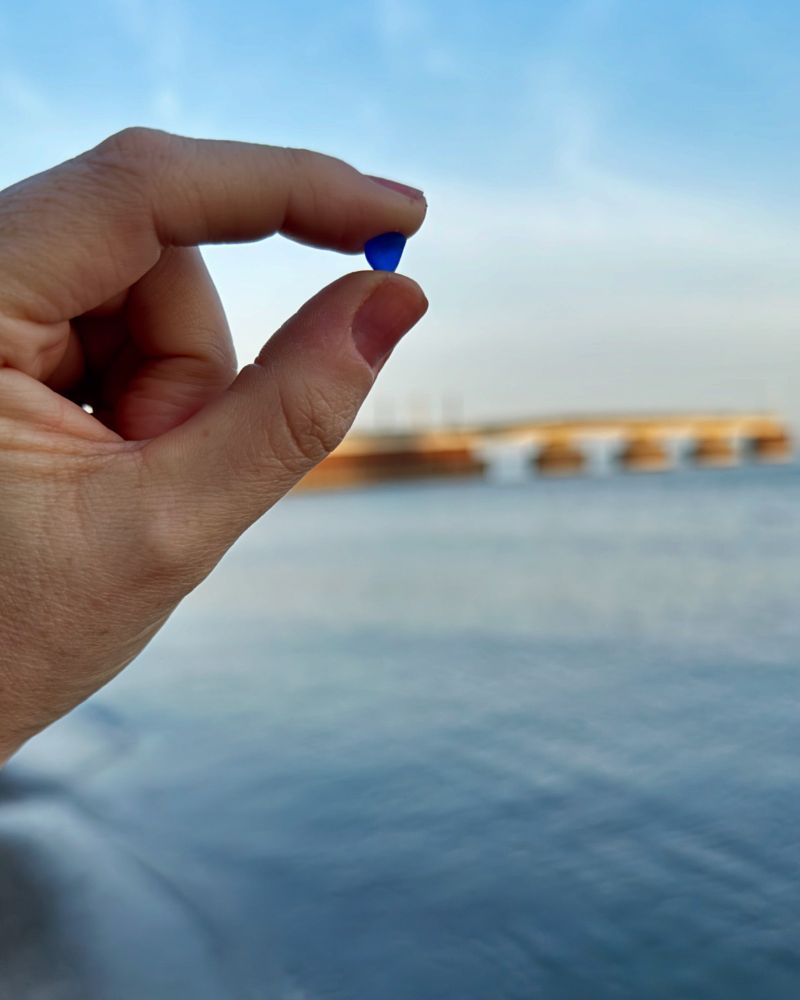 A picture of my hand holding a small piece of blue lake glass in between my thumb and pointer finger. The sky was blue that day. Bradstreet’s Landing pier is out of focus in the background. 