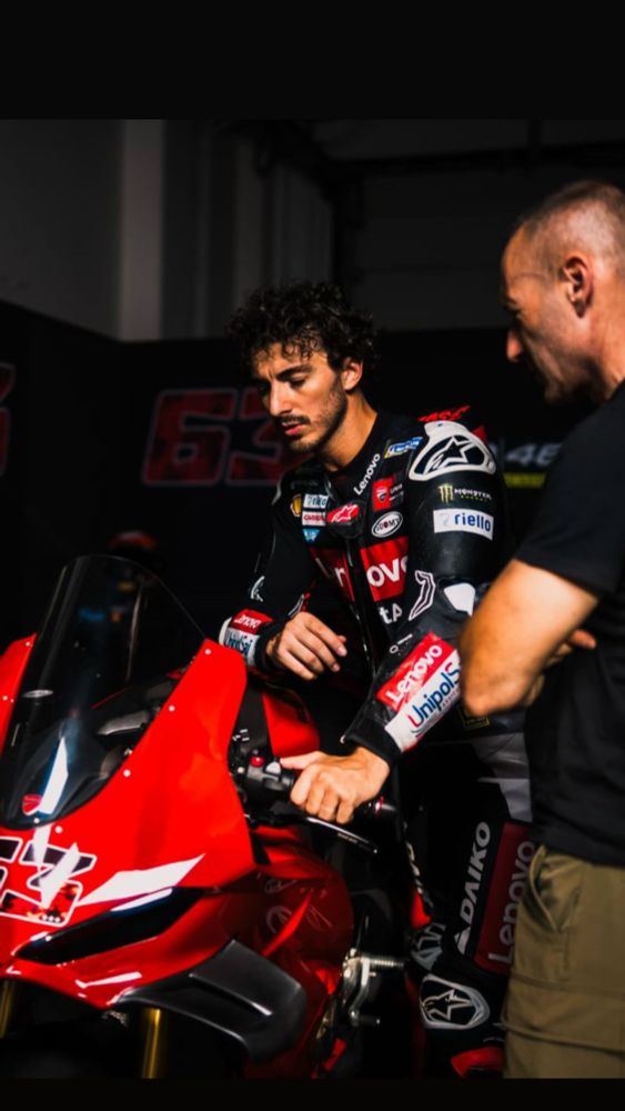 Francesco 'Pecco' Bagnaia on a parked bike, helmet off, speaking with someone