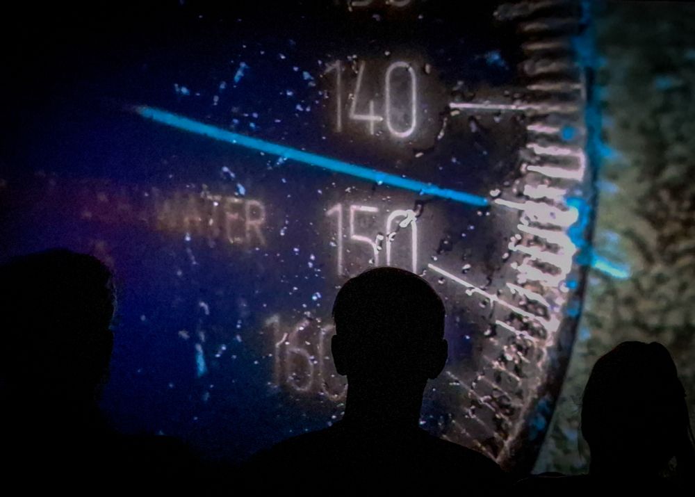 A close up of a dive gauge on a big screen with audience members silhouetted. 