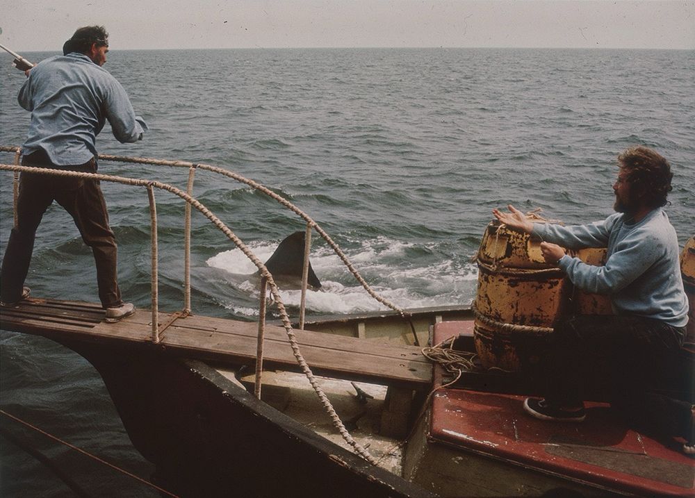 Robert Shaw on the bow of the Orca staring at a shark fin while Richard Dreyfuss ties a tracker to a yellow barrel in a still from Jaws.