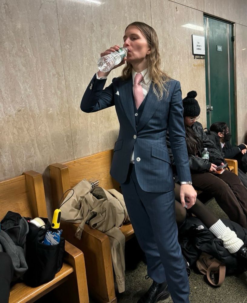 Chelsea Manning in a tailored blue pinstripe suit with a pink tie drinking from a bottle of water alongside a row of benches against a wall.