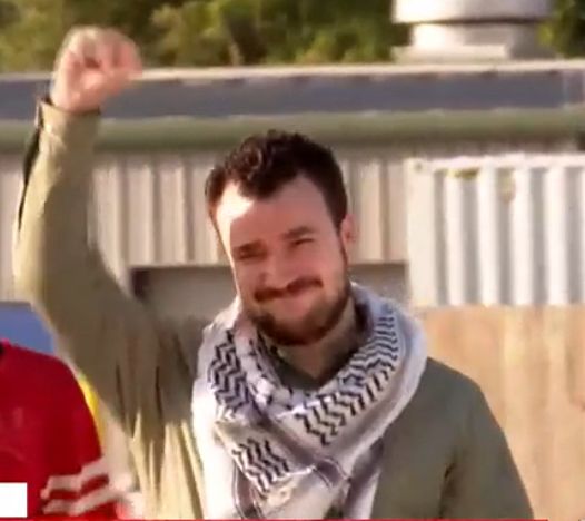 a still of mahmoud khalil as he exited ice detention. he’s wearing a long sleeve khaki button up and a white and black keffiyeh wrapped around his neck. he’s got a big smile on his face and his fist raise in the air, defiant and proud.