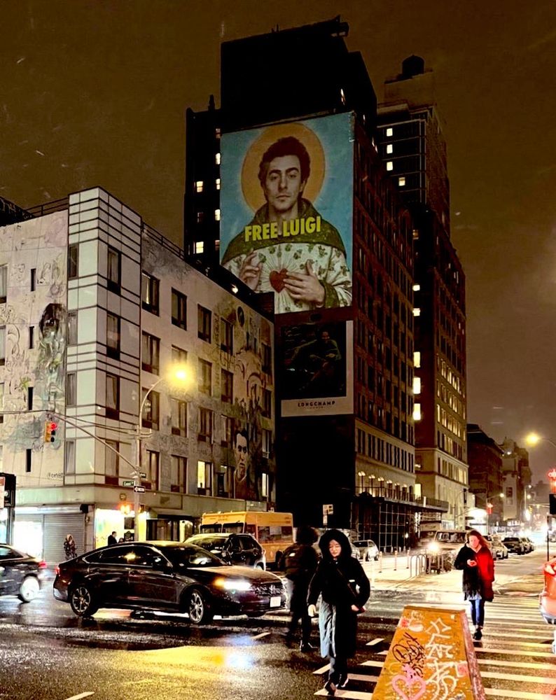 photo of a wintry street, cars and people and snow flurries zooming past. on a building down the block, a massive advertisement hangs overlooking the traffic. it is luigi mangione, edited into the image of a saint. the text ‘free luigi’ is featured prominently in yellow, front and center.