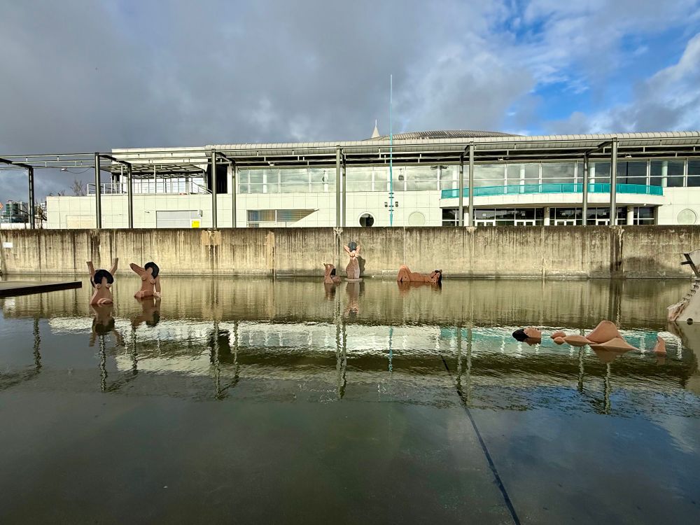 A reflecting pond includes various sculptures of naked women bathing under a cloudy sky. 