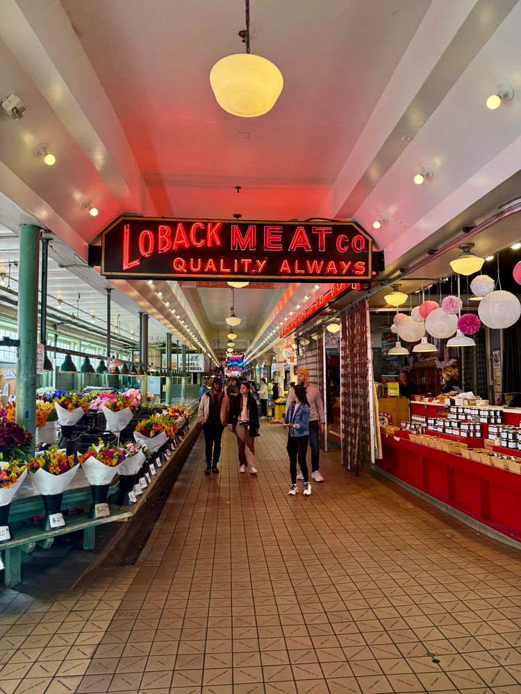 A neon sign reads Loback Meat Co, Quality Always above a walkway lined with flower bouquets
