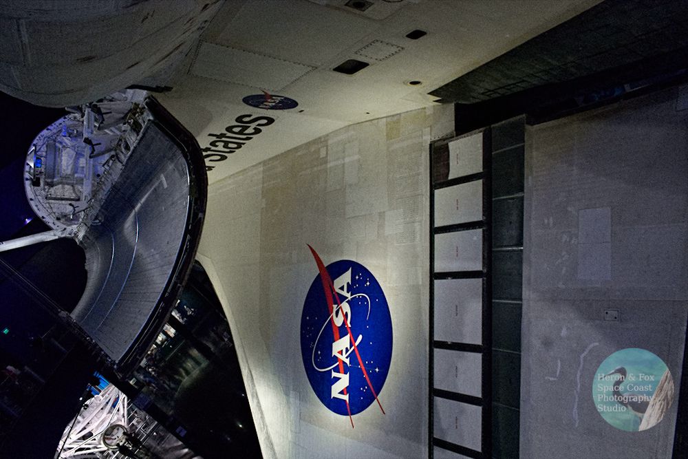An indoor photograph looking down the fuselage of the Space Shuttle Atlantis on its port side, with its payload bay door and wing visible.